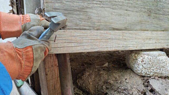 Gloved hands positioning metal nail on wooden beam before hammering
