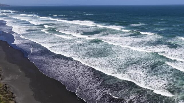 Drone aerial of powerful waves rolling onto black sand coastline of New Zealand's North Island, coastal landscape view.