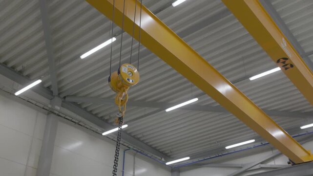 Yellow overhead crane with chain hoist mounted beneath a bright industrial ceiling in a modern factory
