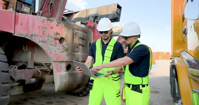 Adult male logistics crew in helmet and hi-vis vest discuss laptop data near crane truck, checking freight schedule, cargo documentation, container yard safety inspection during worksite planning