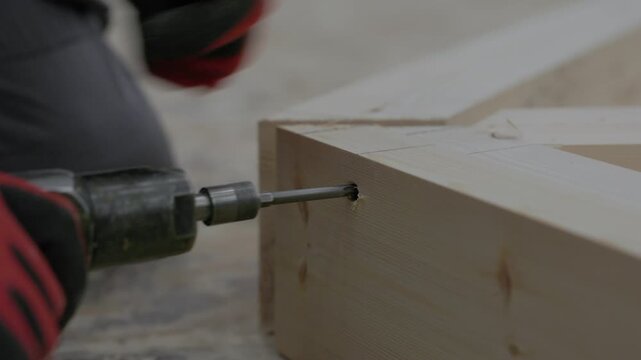 Close-up of a power drill driving a wood screw into a timber beam during frame assembly in a woodworking workshop. Shallow depth of field with copy space