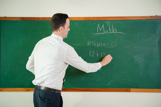 Male teacher writing math equations on a green chalkboard during a classroom lesson. Professional schoolteacher explaining mathematics and writing on chalkboard in elementary school.