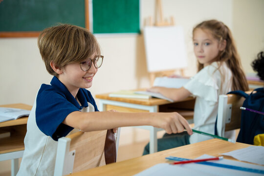 Smiling schoolboy with glasses sharing or passing a pencil to his classmate in classroom. Friendly elementary student sharing stationery with a friend during a lesson in class.