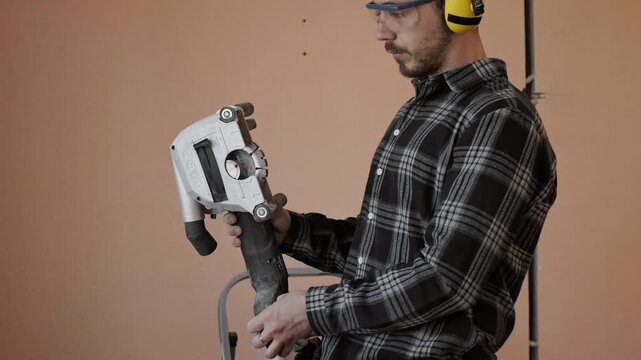 Electrician wearing safety glasses and ear protection holds construction tool and inspects it before electrical work begins