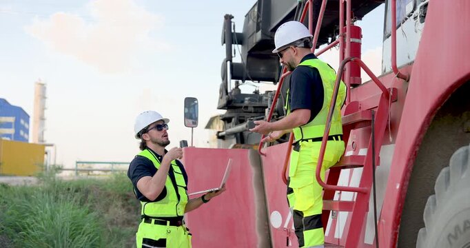 Adult Caucasian male freight supervisor speaks from container truck step while colleague holds laptop, reviewing dispatch data, transport schedule, safety protocol, cargo handling plan at busy yard