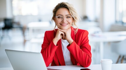 Fototapeta premium Confident female entrepreneur in red jacket and glasses smiling at desk in modern office