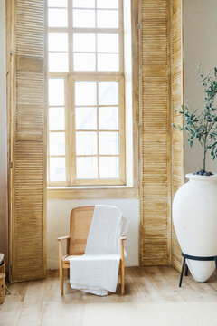 Wooden chair with white blanket near large window and big ceramic vase with plant