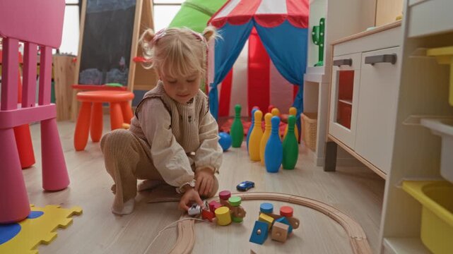 Little blonde girl kneeling, hands on wooden train track and toys on foam playmat in colorful playroom; curiosity playtime memory.