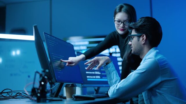 Two asian IT professionals analyze complex data on multiple computer screens in dark, high-tech server room. Diverse male and female team discuss cybersecurity code and business intelligence software