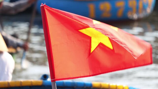 The national flag of Vietnam flutters in the wind, with tourists sailing on round boats in the background, Hoi An Ancient Town, Vietnam