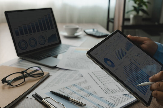 Tablet and laptop display financial dashboards above printed reports on a desk while hands review charts and tables for financial data reporting, analysis, and business performance monitoring.