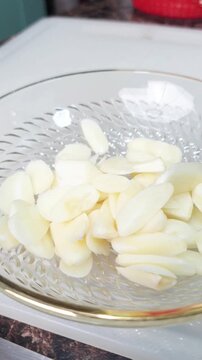 A hand dropping freshly sliced garlic cloves into a clear glass bowl with a gold rim, preparing ingredients for cooking