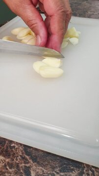 A person using a kitchen knife to slice fresh garlic cloves on a white cutting board for meal preparation