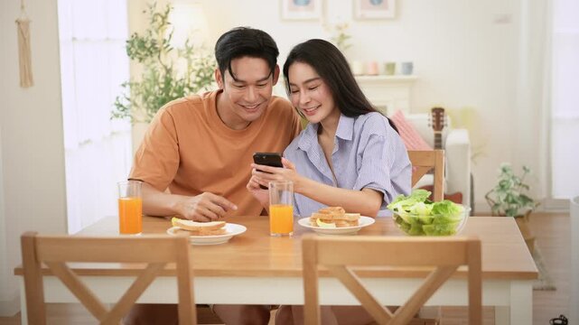 Young Couple Enjoys Breakfast Together While Sharing Laughs and Looking at a Smartphone