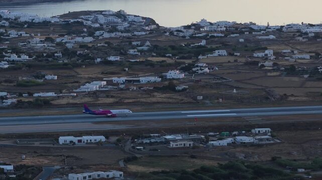Wizz Air airplane landing on Mykonos airport runway during summer vacation in Greece. Cycladic architecture and Aegean Sea from unique tripod elevated perspective