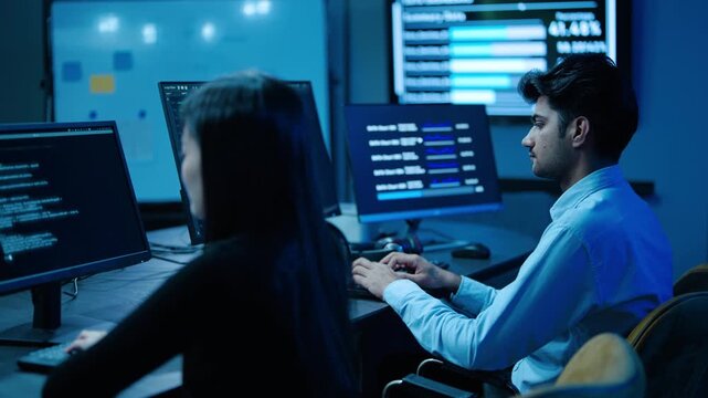 Focused young asian IT professionals work late typing on computer in dark, blue-lit cybersecurity control room. They actively analyze complex data networks and write software code on glowing monitors