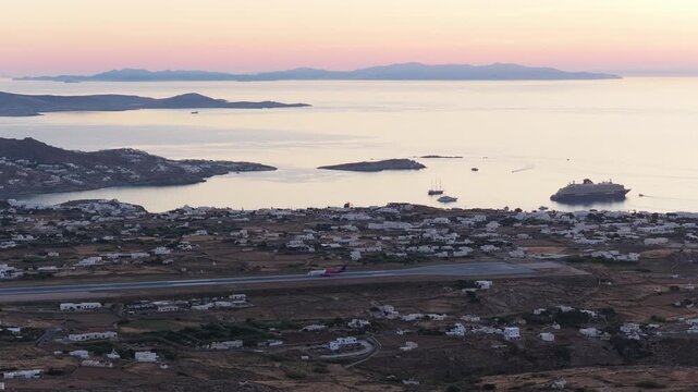 Commercial passenger airplane taxiing the runway in Mykonos Island National Airport at sunset with a cruise ship anchored in the Aegean Sea and other Cyclades islands in background