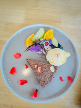Top view of a rich chocolate brownie served with vanilla ice cream, fresh fruits, whipped cream, blueberries, edible flowers, and rose petals on a ceramic plate. A beautifully presented gourmet desser