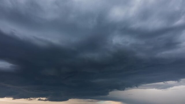 Dark storm cloud shelf lowering over low horizon, advancing across wide sky with dramatic layered undersides, scalloped base, and heavy moody atmosphere above distant land