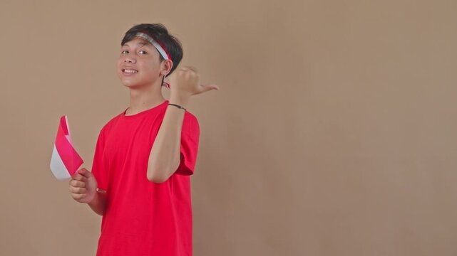 Happy Indonesian teen holding mini flag and pointing his thumb back at copy space 
