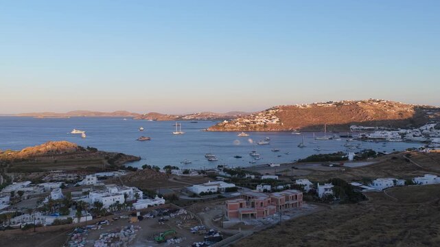 Golden hour light illuminating Ornos bay in Mykonos, Greece, numerous sailing boats and luxury yachts anchor near charming white houses built on arid coastal hills
