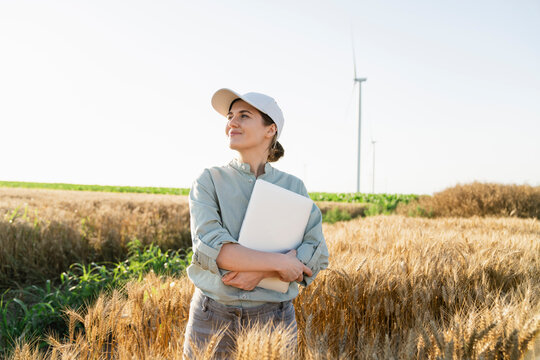 Smiling farmer wearing cap and holding laptop at wheat field
