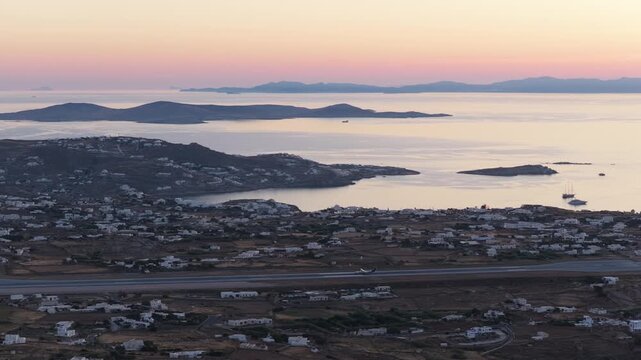 Commercial jetliner taking off at Mykonos Airport in Greece, overlooking Aegean Sea, featuring distant islands and pink sunset sky during summer holiday