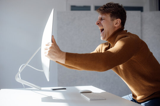 Angry businessman lifting computer monitor screaming at desk in office