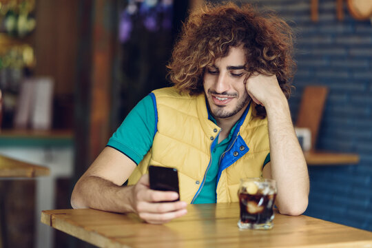 Smiling young man with beard and curly hair looking at cell phone in a pub
