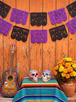 Hand-painted guitar and skull-themed paper banners hang on a weathered orange wall, surrounded by marigold flowers, candles, and colorful sugar skull decorations for a festive celebration.