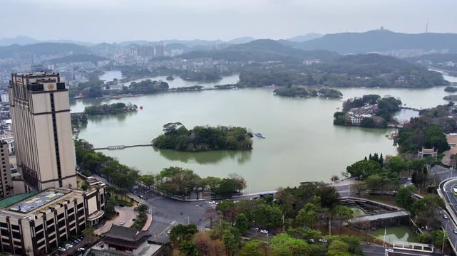 This is an aerial view of a large urban lake with tree-covered islands, adjacent buildings and roads, and distant hazy hills.