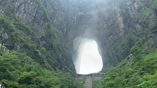 Tourists climbing steep staircase toward massive natural limestone arch in mountain. Tianmen mountain heaven gate cave with lush green forest. Scenic Asian landmark and outdoor travel adventure.
