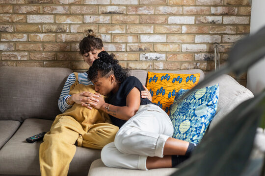 Young woman kissing pregnant girlfriend stomach while sitting on sofa at home