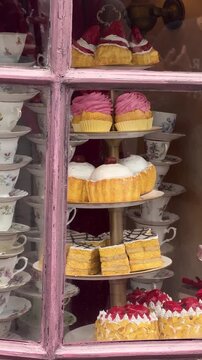Assorted cakes and muffins displayed on a tiered stand in a shop window. Vintage floral teacups stacked beside delicious pastries. Bakery showcase for dessert cafe business concept.
