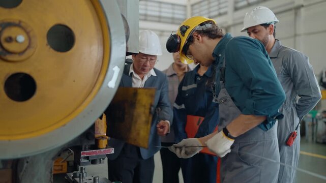 Factory workers inspect heavy machinery with a supervisor. They wear protective gear and gloves, focused on their task. The team discusses plans using a tablet and clipboard