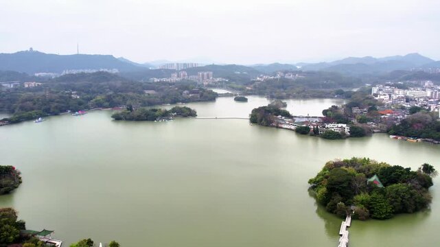 An aerial perspective shows a large lake dotted with tree-covered islands, bordered by hills and mixed settlements under an overcast sky.