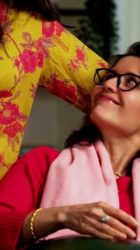 Closeup portrait of a happy Indian Asian pretty young girl in her twenties and her mother in fifties, both looking at the camera while sitting close together on a cozy sofa at home, enjoying their tim