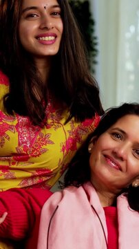 Closeup portrait of a happy Indian Asian pretty young girl in her twenties and her mother in fifties, both looking at the camera while sitting close together on a cozy sofa at home, enjoying their tim