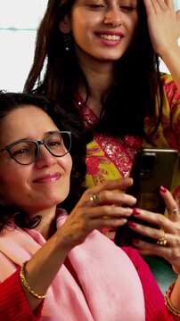 Indian Asian pretty young girl in her twenties and her mother in fifties sitting together on a sofa at home, both using a smartphone, enjoying cozy time and connection in a comfortable living room