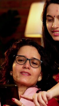 Indian Asian pretty young girl in her twenties and her mother in fifties sitting together on a sofa at home, both using a smartphone, enjoying cozy time and connection in a comfortable living room