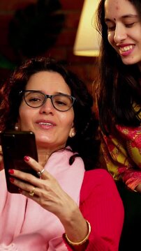 Indian Asian pretty young girl in her twenties and her mother in fifties sitting together on a sofa at home, both using a smartphone, enjoying cozy time and connection in a comfortable living room
