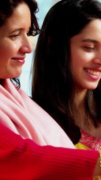 Indian Asian pretty young girl in her twenties and her mother in fifties sitting together on a sofa at home, both using a smartphone, enjoying cozy time and connection in a comfortable living room