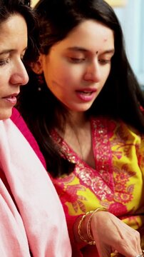 Indian Asian pretty young girl in her twenties and her mother in fifties sitting on a sofa at home, using a laptop to watch movies, surf the internet, shop online, or make video calls