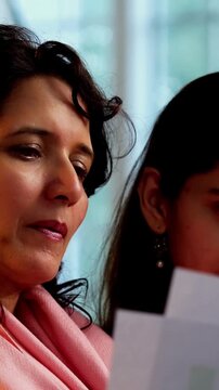 Indian Asian mother in her fifties and her young daughter working together on budgeting and accounting with a laptop and account statements while sitting on a sofa at home