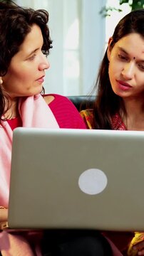 Indian Asian pretty young girl in her twenties and her mother in fifties sitting on a sofa at home, using a laptop to watch movies, surf the internet, shop online, or make video calls