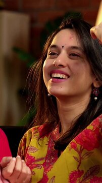Indian Asian mother in her fifties with her pretty young daughter, both sitting on a cozy sofa at home, watching TV together with a remote in hand