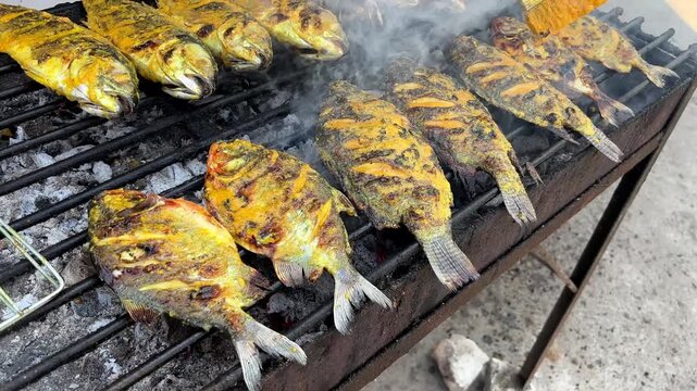 Grilling whole fresh sea fish on an open charcoal grill while brushing traditional yellow turmeric spice paste at a street food