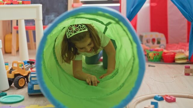 Girl crawling through a bright green play tunnel, hands and knees on floor, wearing plastic tiara and jeans in building; joyful play.