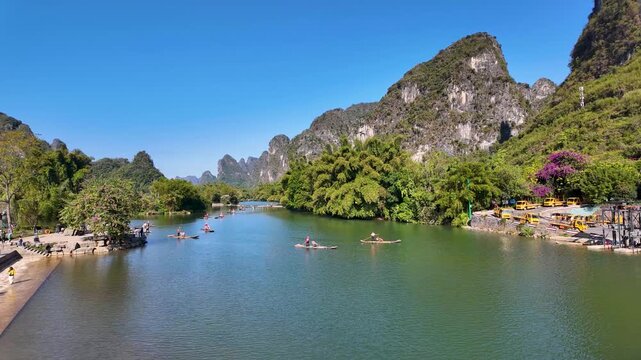 A calm river winds past limestone karsts and lush vegetation, with small boats and people on the water and shore under a clear blue sky.
