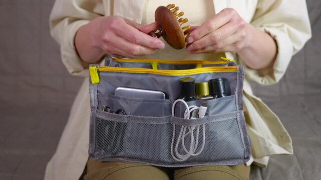 Woman organizing travel bag putting accessories inside. Female hands carefully placing a wooden comb into a gray travel organizer bag filled with cosmetics, cables, and a power bank